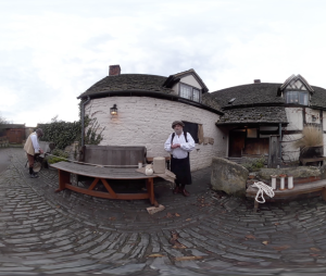 A person in historical clothing stands by a wooden table outside a stone cottage with a tiled roof. Another person is seen walking away on a cobbled path. Rope and jars are on the table.