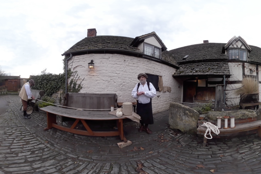 A person in historical clothing stands by a wooden table outside a stone cottage with a tiled roof. Another person is seen walking away on a cobbled path. Rope and jars are on the table.
