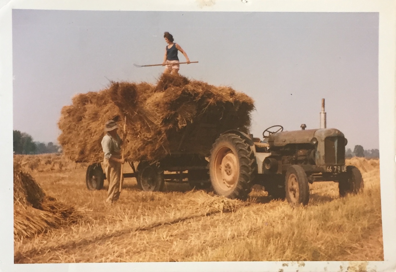 A person stands on a hay waggon stacked with straw, holding a tool, while another stands beside it. An old tractor is attached, and the scene is set in a sunlit field.