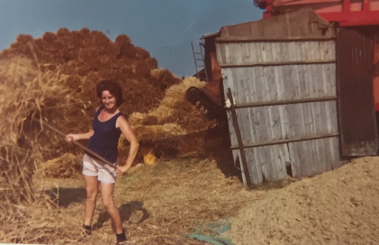 A woman in a blue vest and white shorts is smiling whilst holding a pitchfork in front of haystacks and a rustic wooden shed on a sunny day.