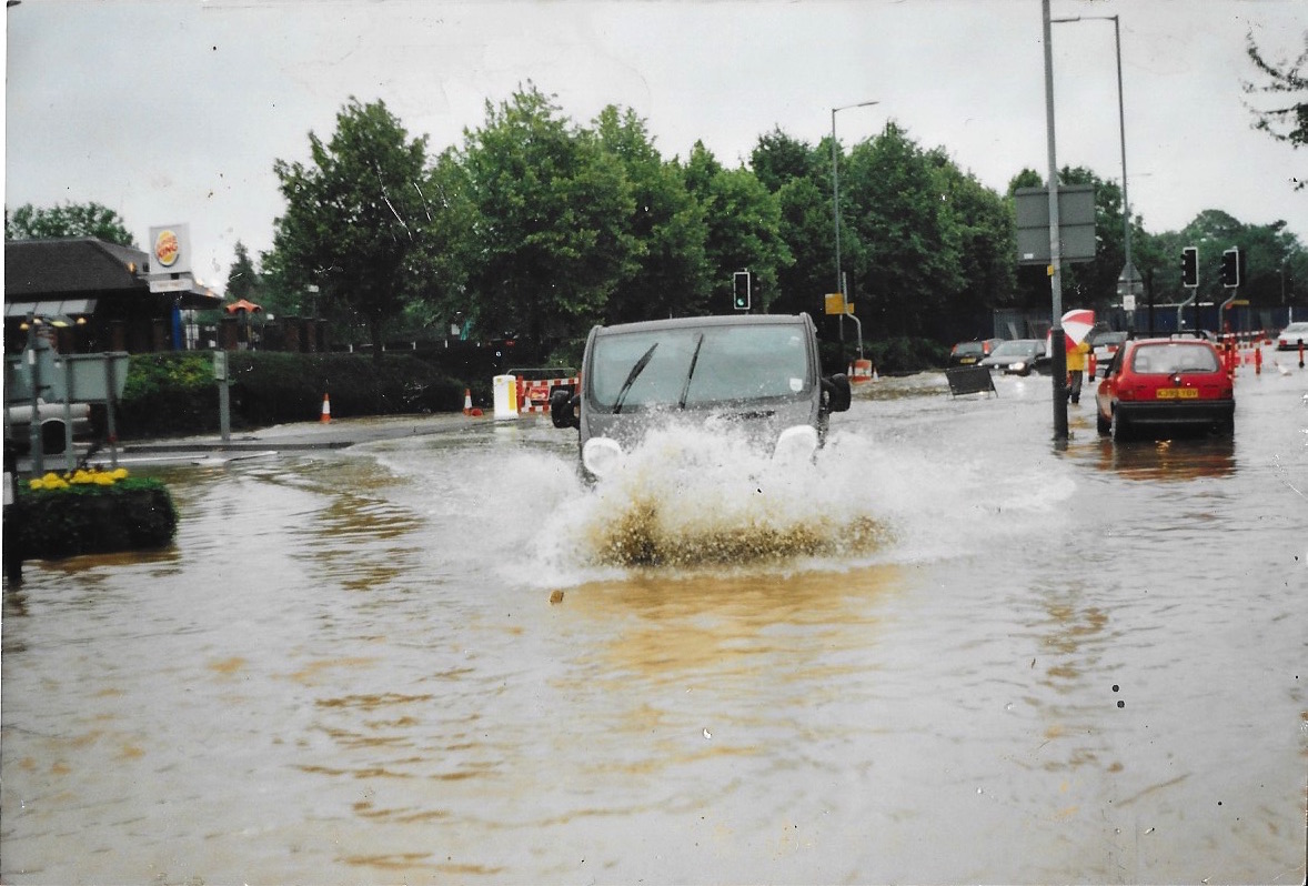 A grey vehicle drives through a flooded street, creating a large splash. Other cars and traffic lights are visible, with trees and buildings in the background.