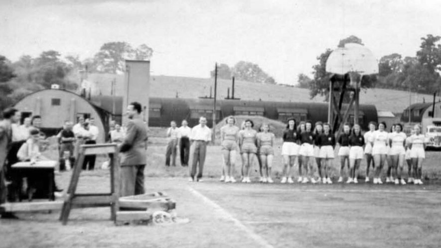 A vintage black-and-white photo shows women's basketball teams lined up outdoors, with men and officials standing nearby. Industrial buildings and a basketball hoop are visible in the background.