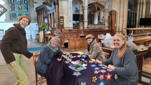 Four women smile while crafting colourful paper stars at a table inside a church. One stands, three are seated, surrounded by art supplies and stained glass windows in the background.