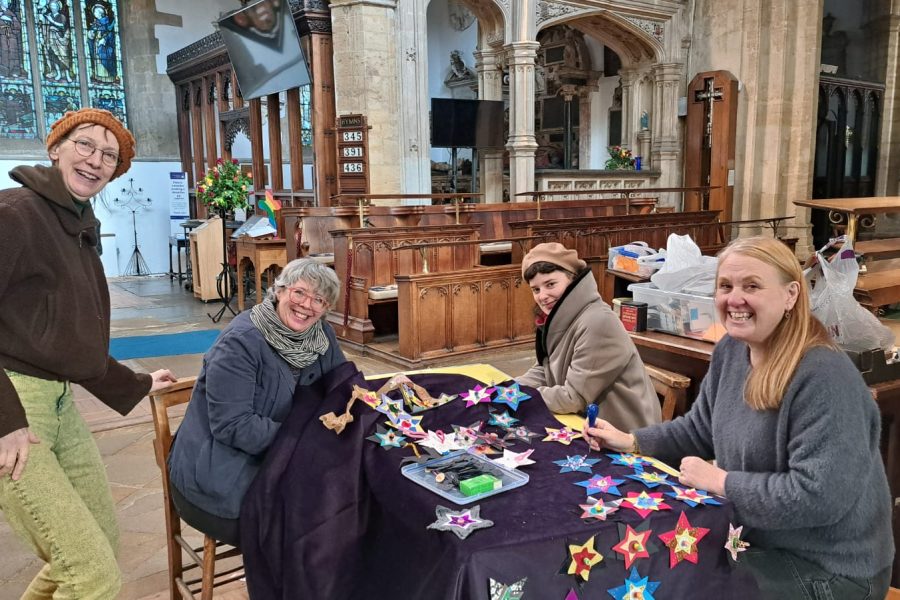 Four women smile while crafting colourful paper stars at a table inside a church. One stands, three are seated, surrounded by art supplies and stained glass windows in the background.