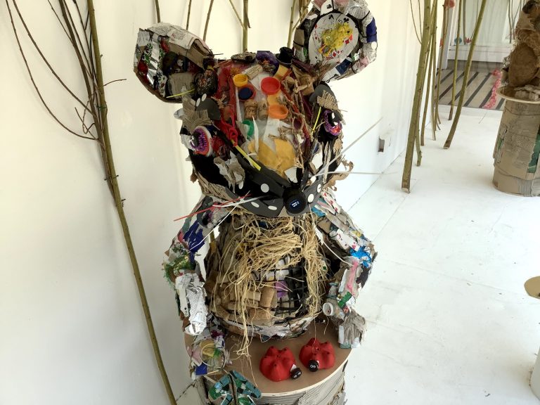 A large mouse sculpture made from recycled materials like cardboard, plastic, and paper stands on a white floor, with two small red objects at its feet and tall sticks in the background.
