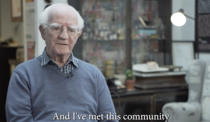 An elderly man with white hair and glasses sits indoors, wearing a blue jumper over a collared shirt. He is speaking, with the caption: And I've met this community. The background shows shelves and a lamp.