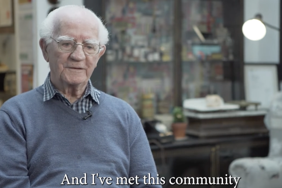 An elderly man with white hair and glasses sits indoors, wearing a blue jumper over a collared shirt. He is speaking, with the caption: And I've met this community. The background shows shelves and a lamp.